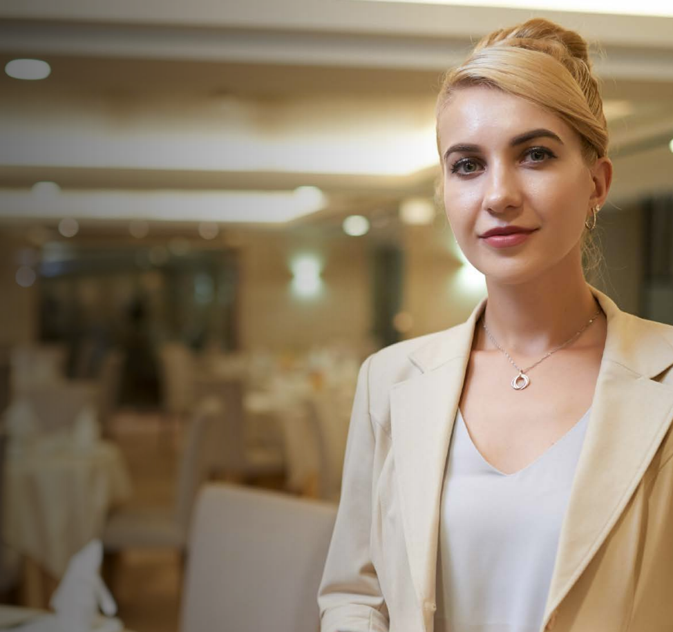 Woman with arms crossed, wearing blue polka dot shirt, standing outside a building, looking directly at the camera.