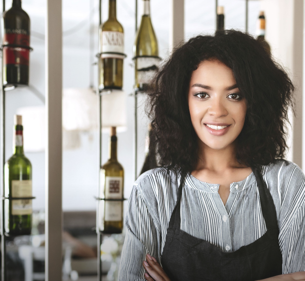 Woman with arms crossed, wearing blue polka dot shirt, standing outside a building, looking directly at the camera.