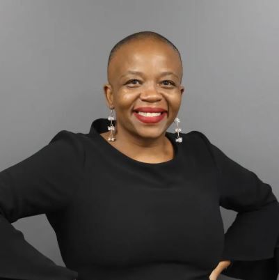 Smiling woman with shaved head, wearing a black top and dangling earrings, against a gray background. Tumi Nkosi