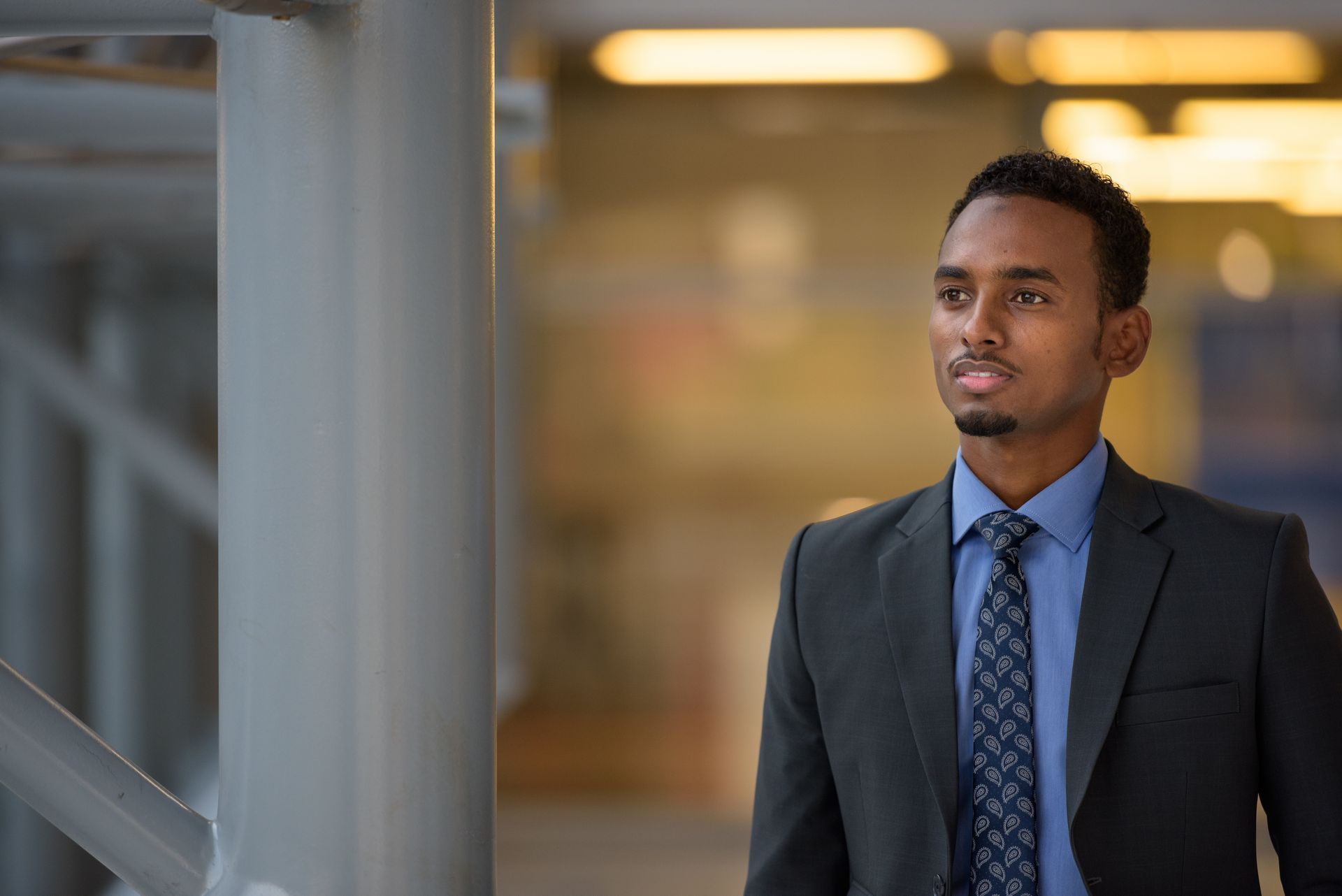 Two people in navy suits, standing in a well-lit office setting. The woman in the front is smiling, hands clasped.