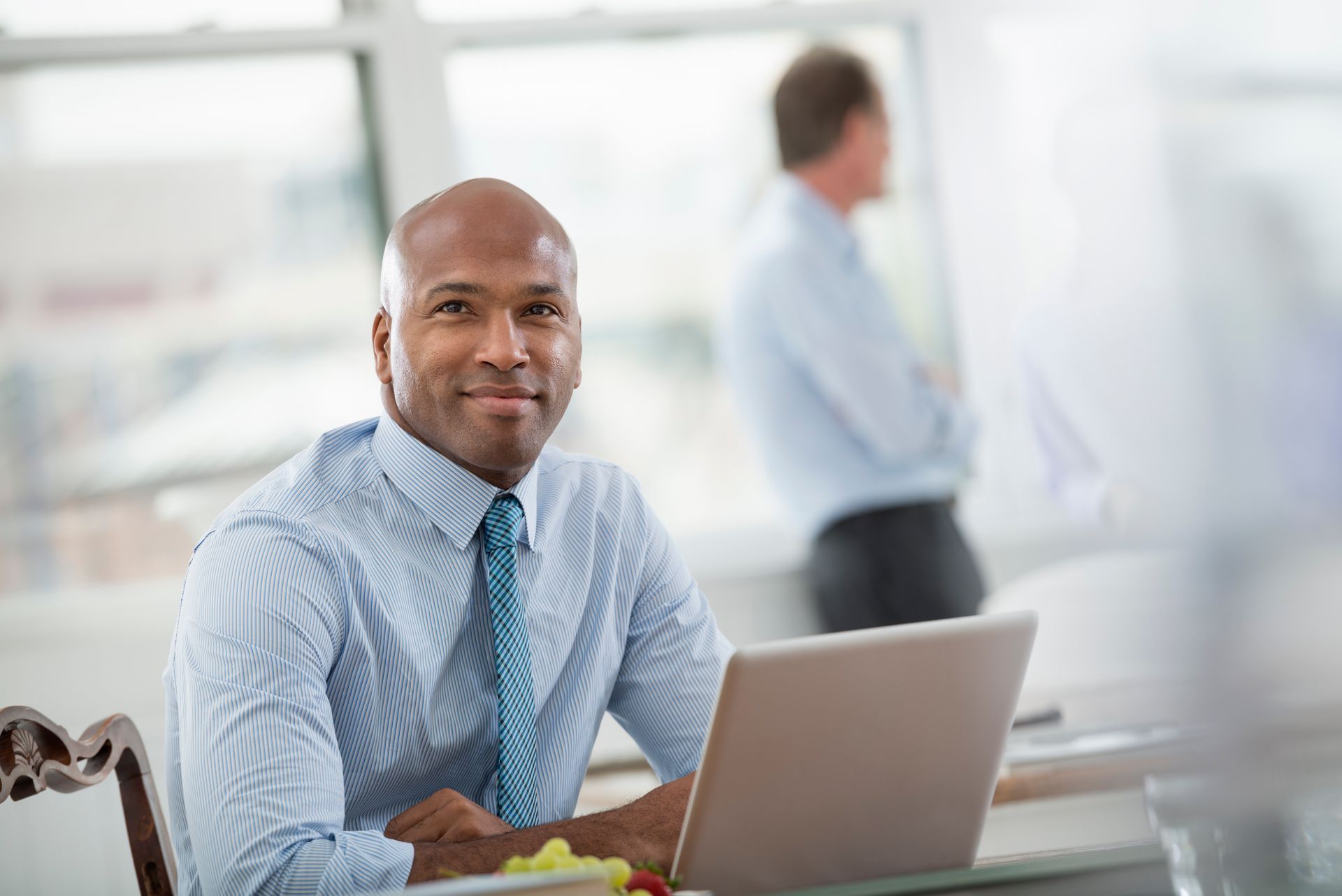 Man in blue striped shirt, smiling at the camera, working on laptop at a desk; another man in the background.