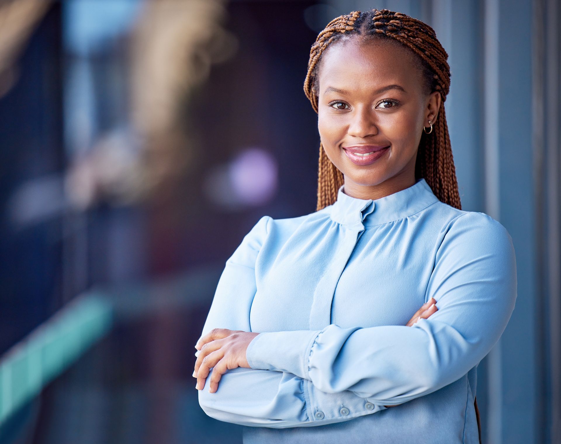 Woman with arms crossed, wearing blue polka dot shirt, standing outside a building, looking directly at the camera.