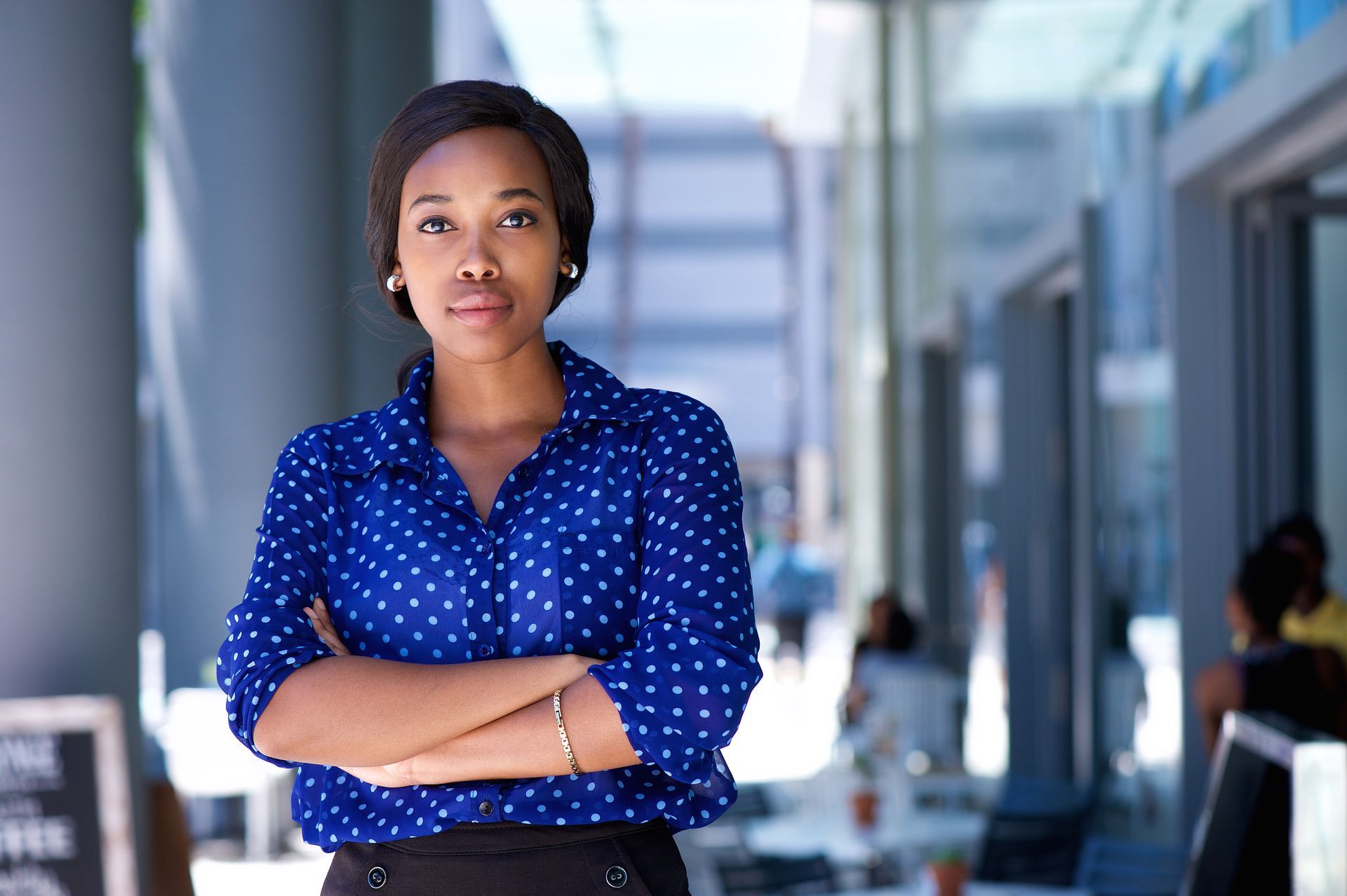 Woman with arms crossed, wearing blue polka dot shirt, standing outside a building, looking directly at the camera.