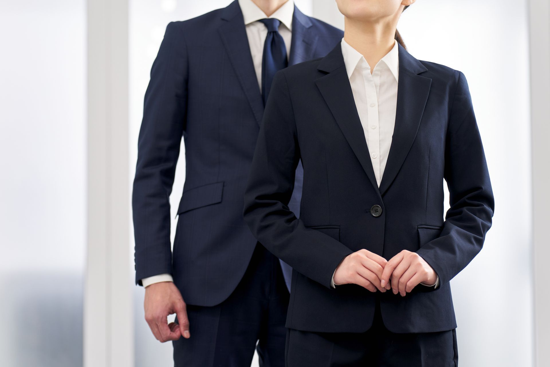 Two people in navy suits, standing in a well-lit office setting. The woman in the front is smiling, hands clasped.