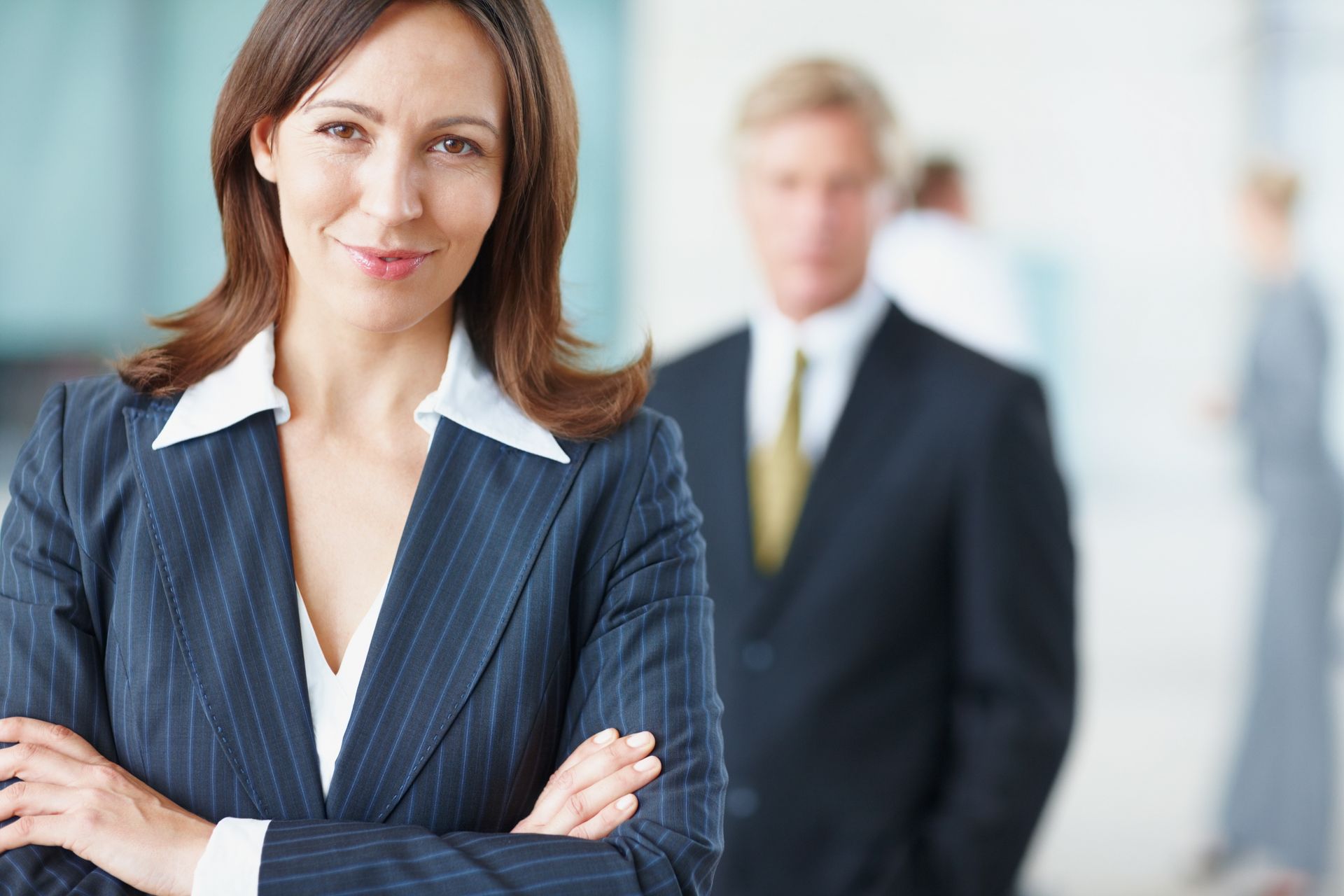 Woman in business suit with arms crossed, smiling, with colleagues in the background.