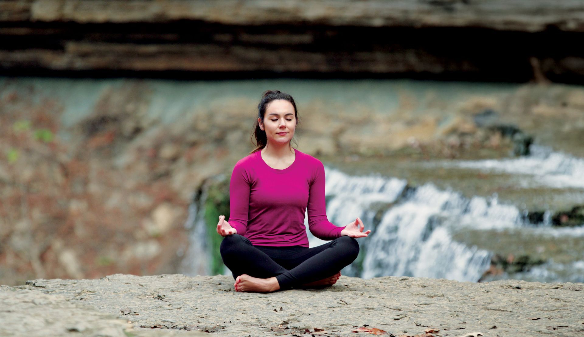 A person meditates in a cross-legged seated pose on a rocky ledge in front of a cascading waterfall.