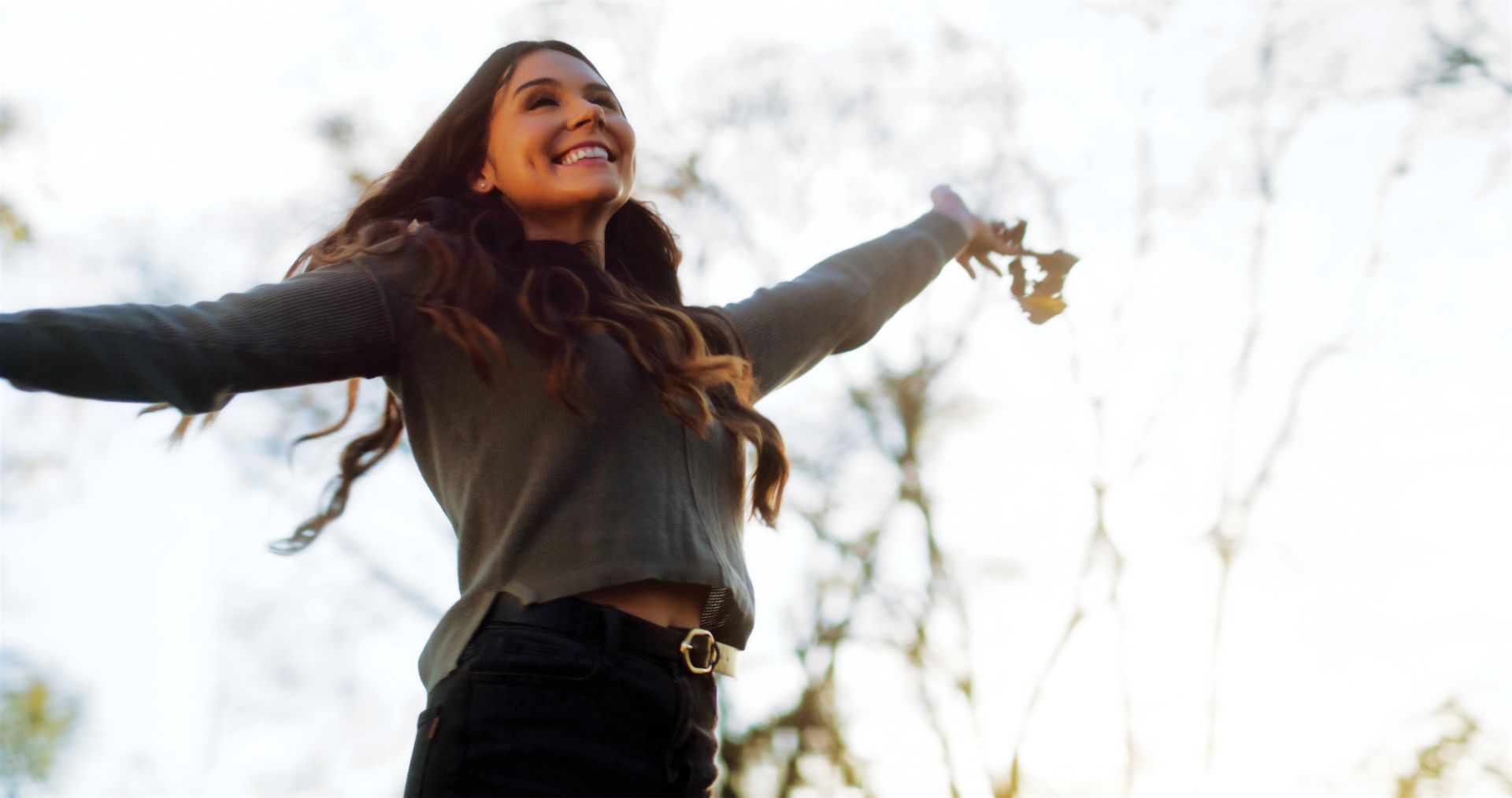A person standing outdoors with arms outstretched, smiling, and holding a dry leaf against a bright, sunlit background.