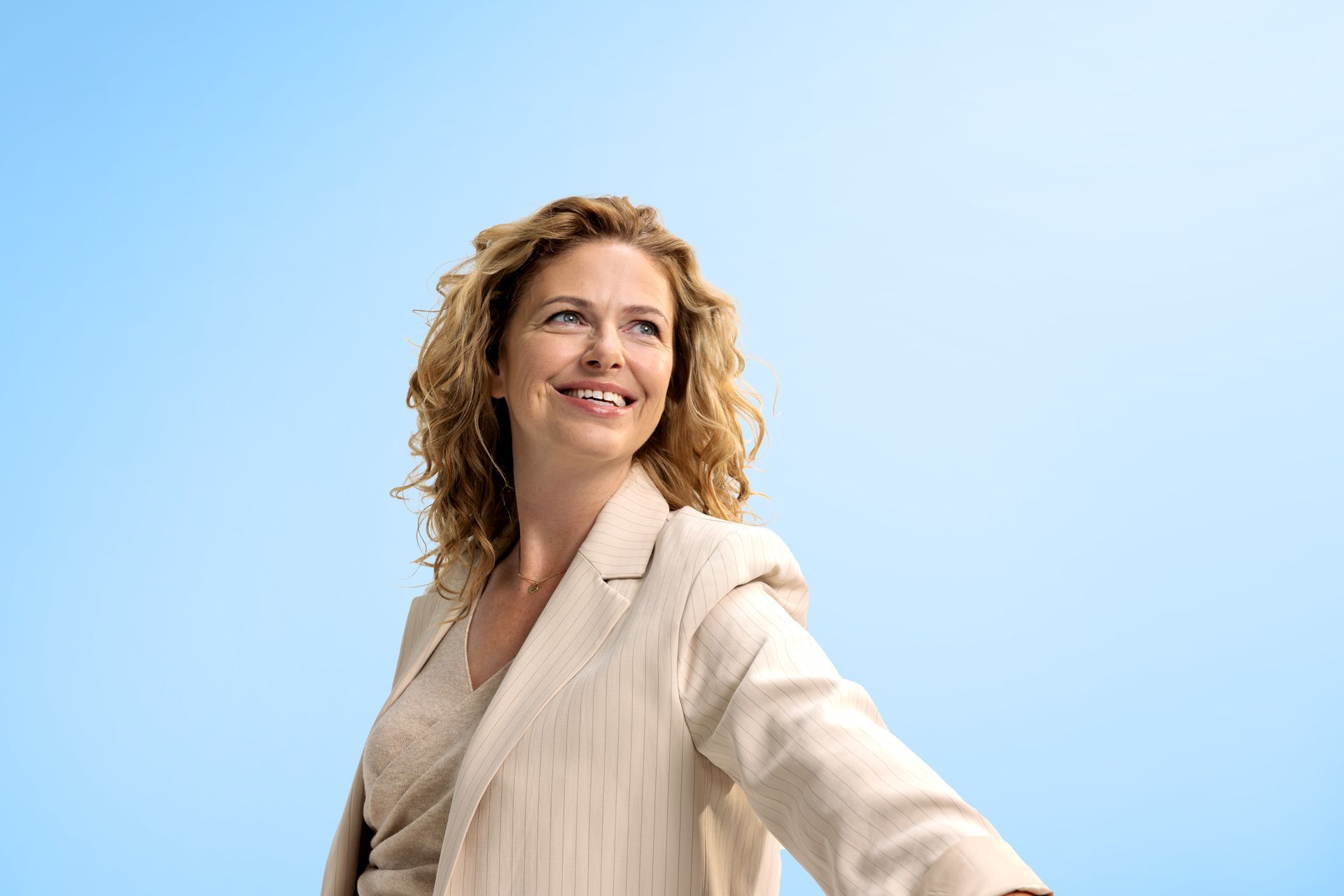 A smiling person with curly hair wears a beige blazer, looking off to the side against a light blue background.