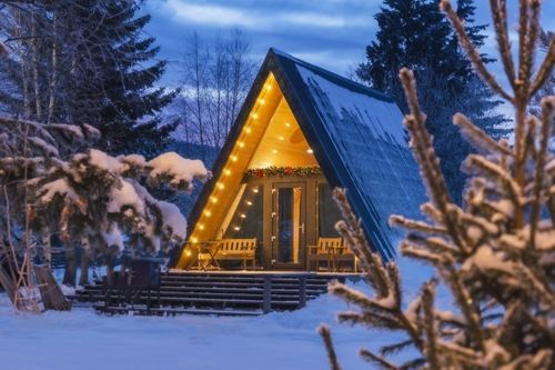 A cozy A-frame cabin surrounded by snow-covered trees at dusk, warmly lit with string lights,