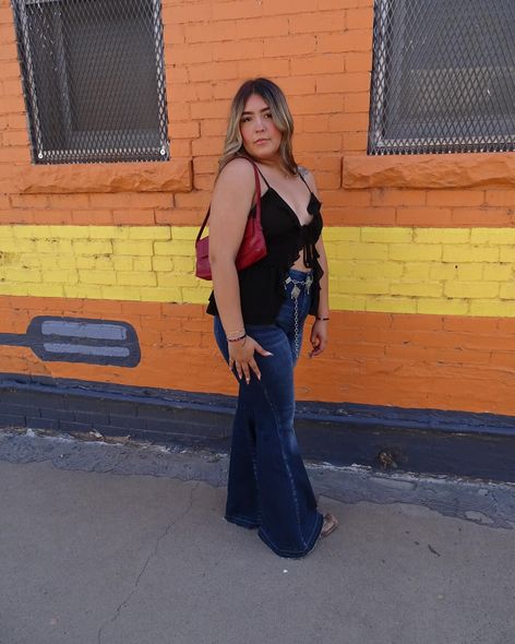 A person stands in profile against an orange and yellow brick wall, wearing a black top, dark flare jeans, and a red bag.