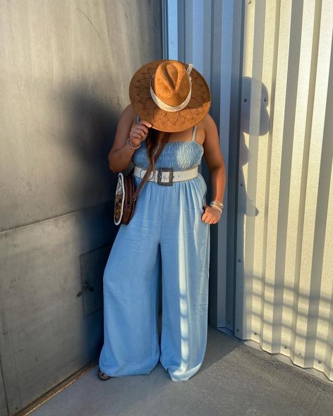 A person wearing a wide-brimmed brown hat, a blue denim jumpsuit, and a wide white belt stands against a metal wall.