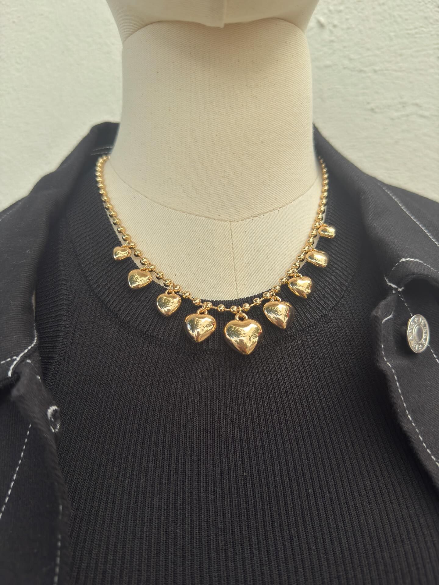 Gold heart charm necklace on a beige mannequin wearing a black top and jacket