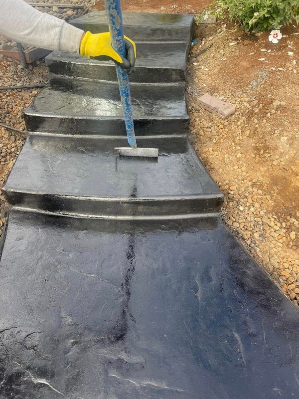 Person with yellow gloves applying black sealant to outdoor concrete steps with a long-handled brush.