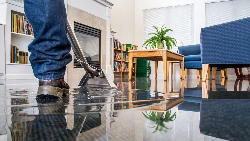 A man is using a vacuum cleaner to clean a flooded floor in a living room.