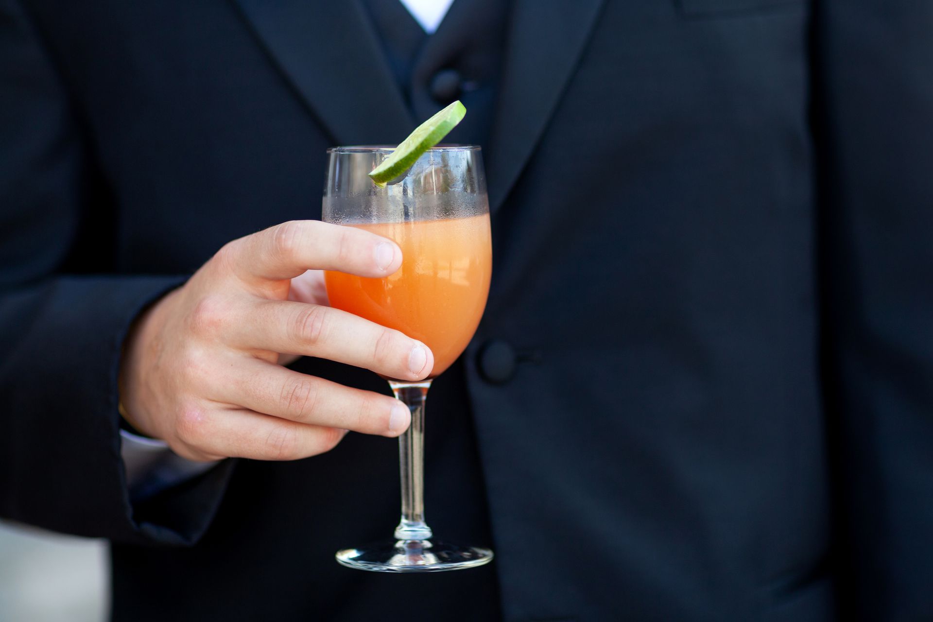 Man in black suit holding a cocktail in a stemmed glass, orange drink with a cucumber garnish.