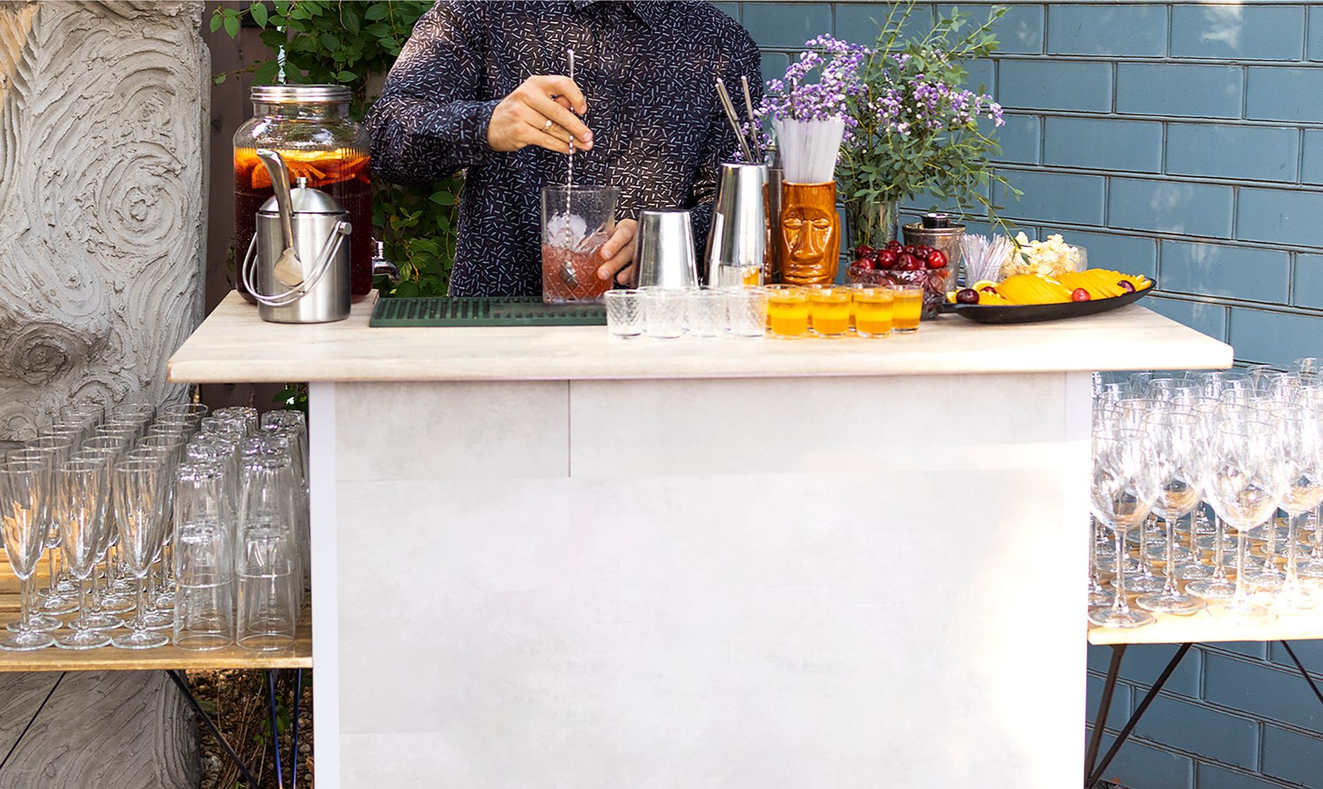 Bartender mixing a drink at an outdoor bar. Drinks, flowers, and fruit on the bar.