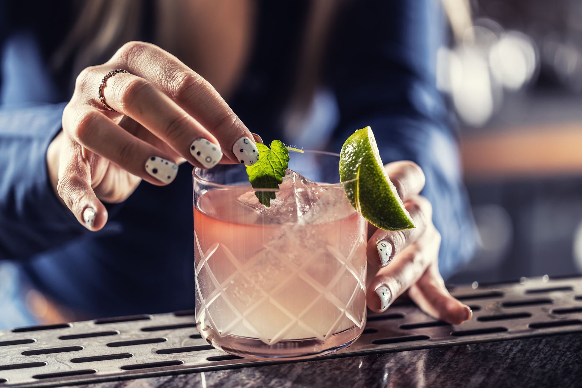 Bartender garnishes cocktail with mint and lime. Pink drink in diamond-patterned glass, on bar.
