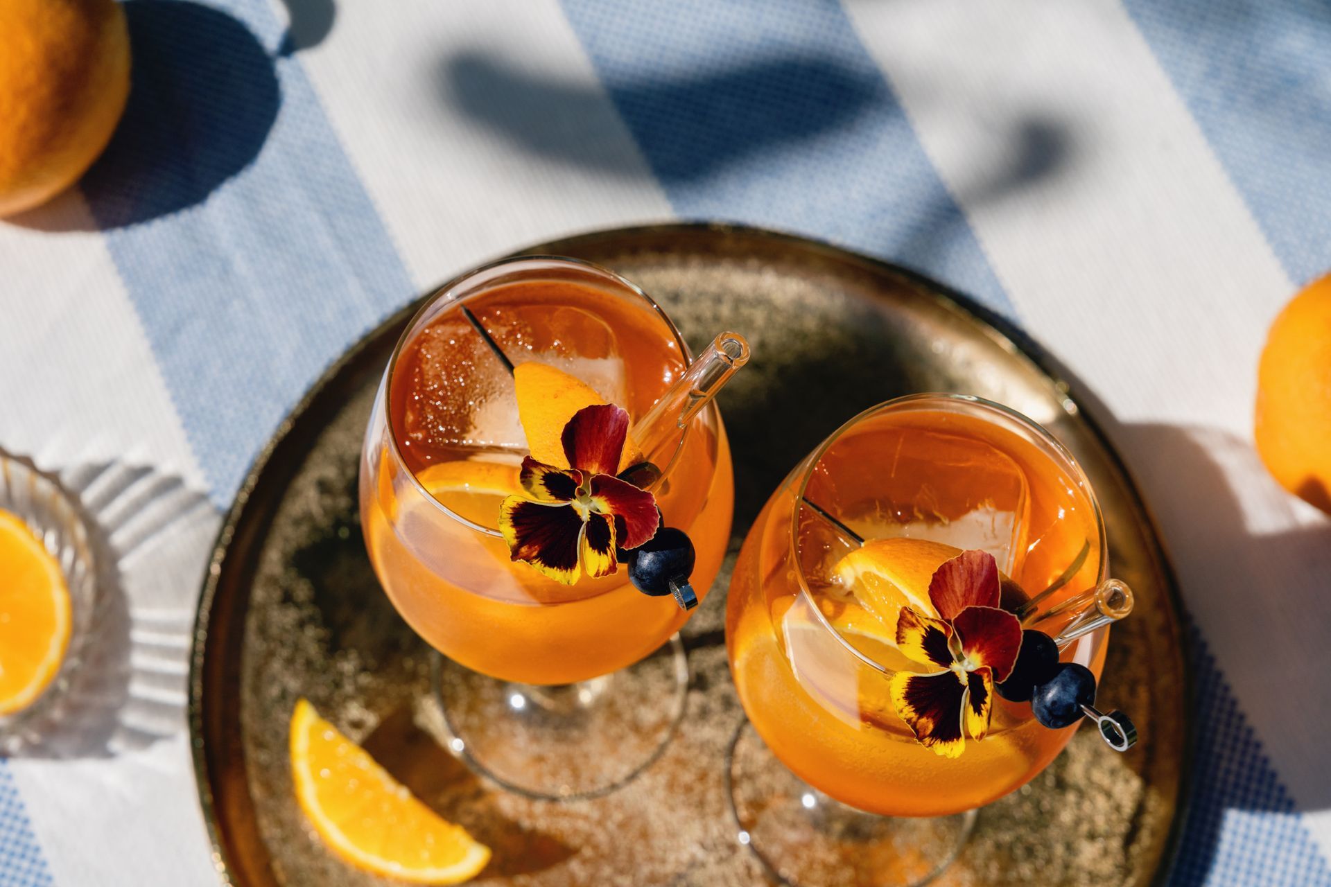 Two orange cocktails with floral garnishes on a tray, oranges, and a striped tablecloth.
