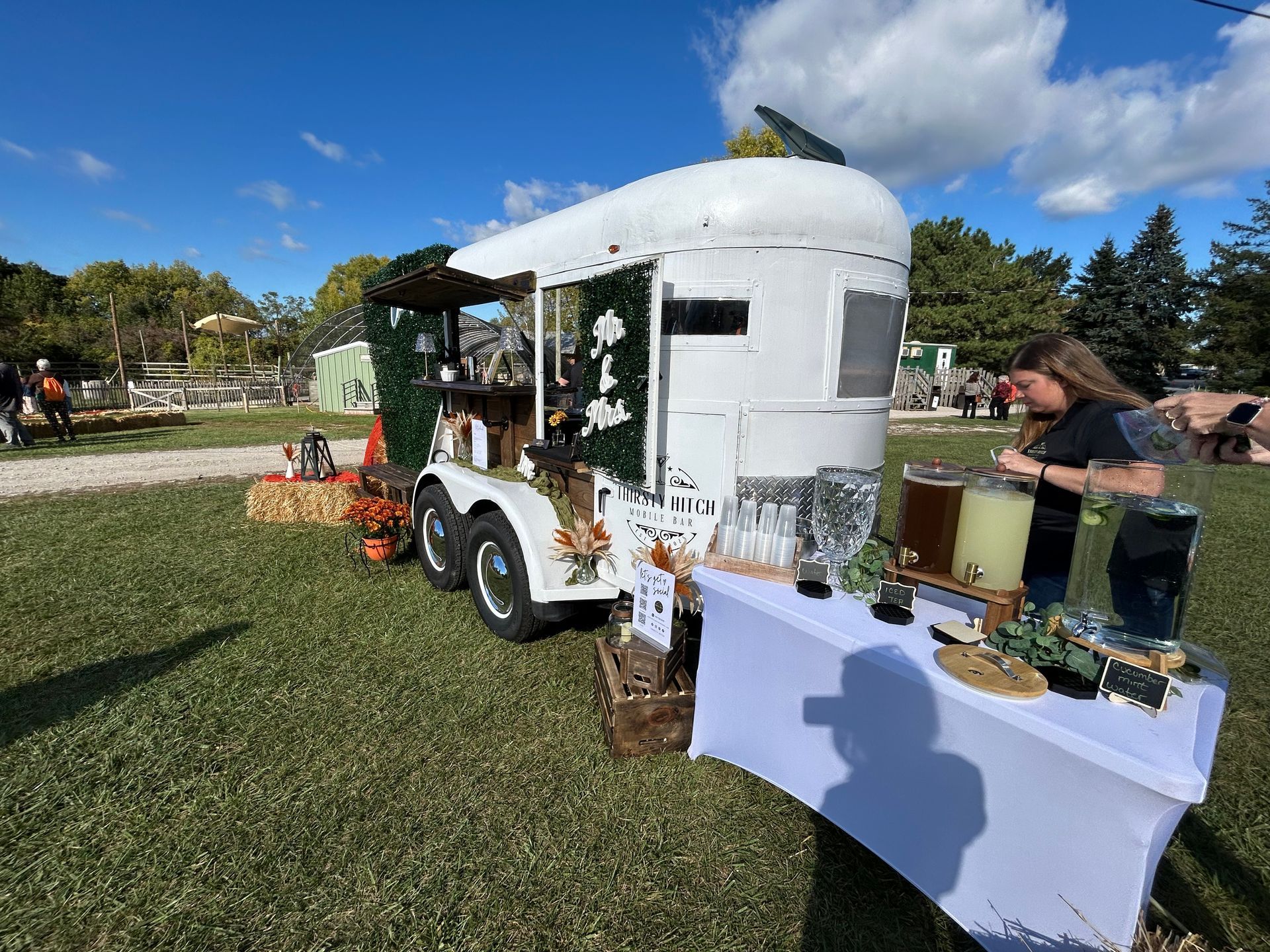 White horse trailer converted into a bar, person serving drinks at an outdoor event.