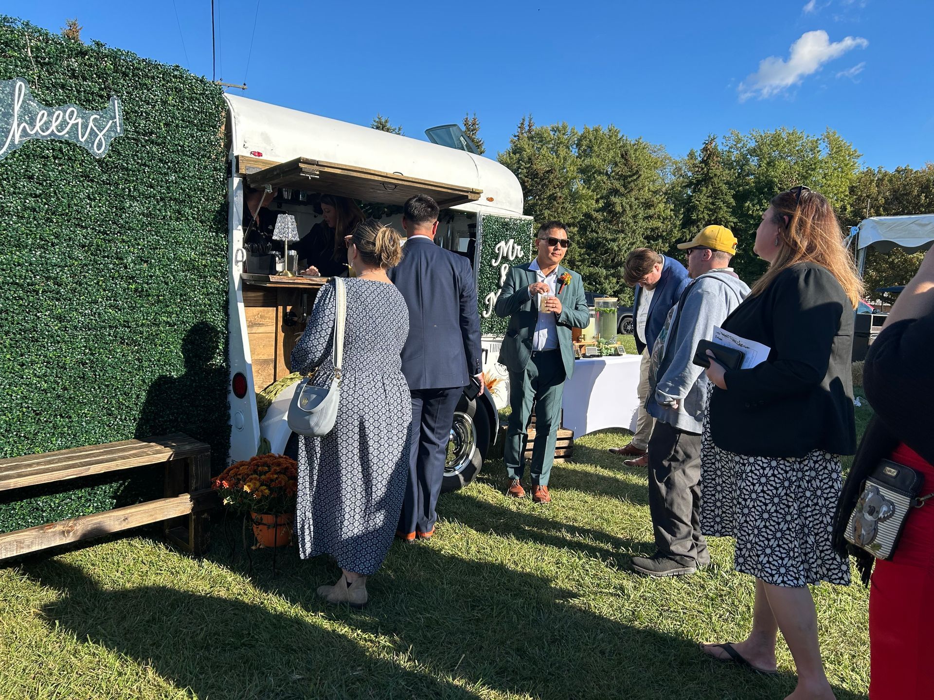 People wait in line at a mobile bar decorated with greenery, set on a grassy area with a blue sky.