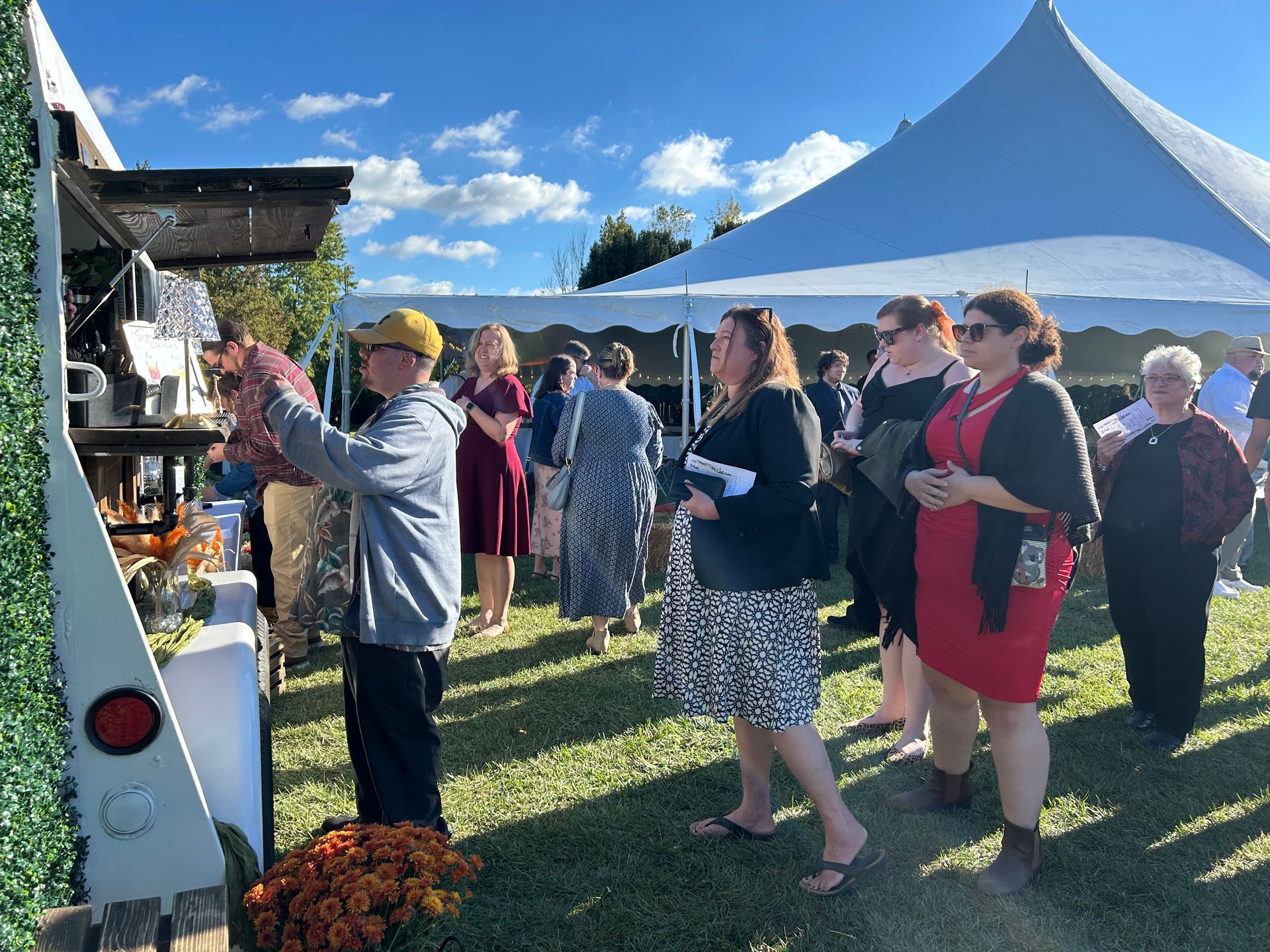 People lined up at an outdoor food/beverage station, with a tent in the background.