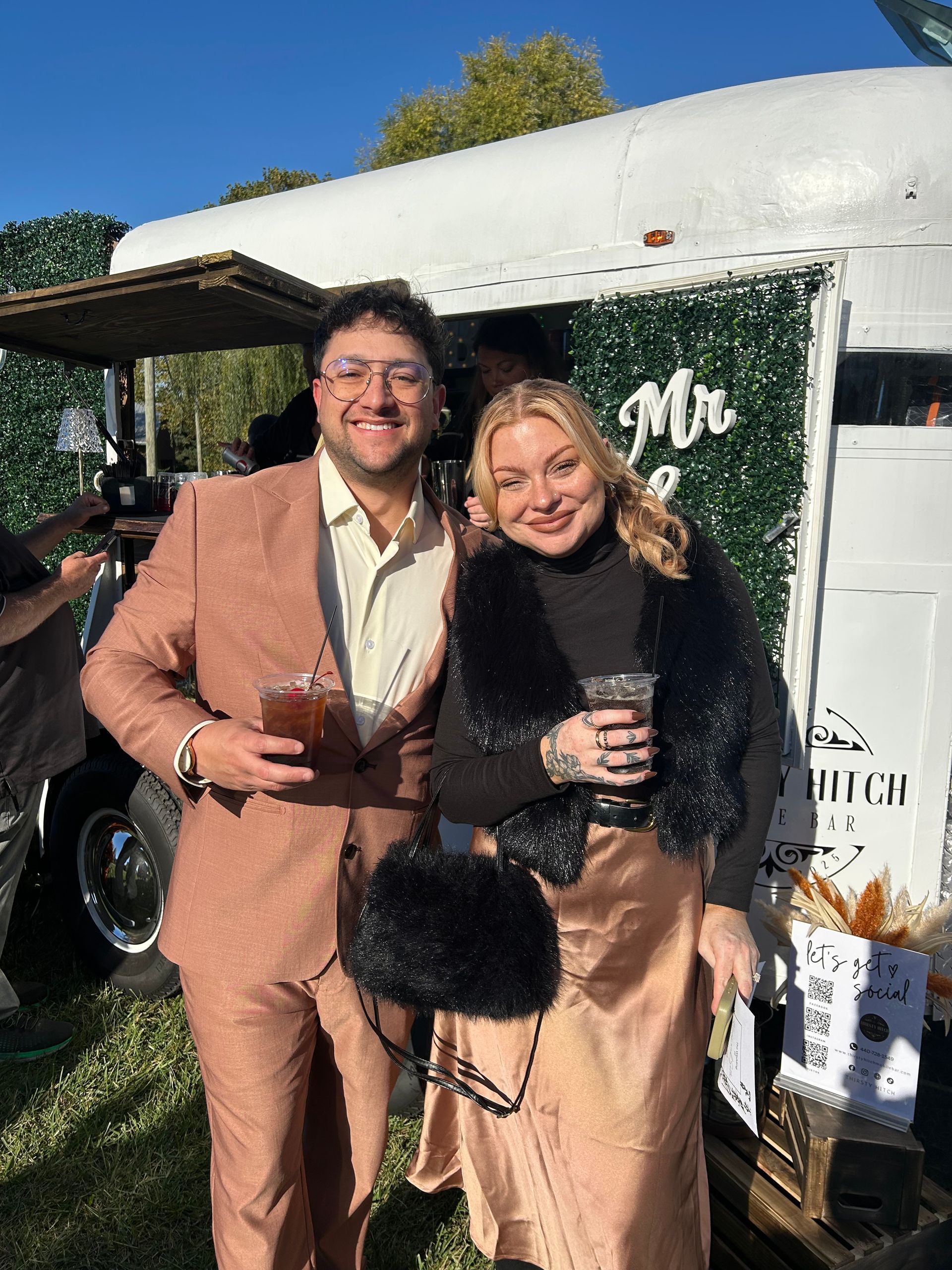 A smiling couple in formal attire stands in front of a mobile bar.