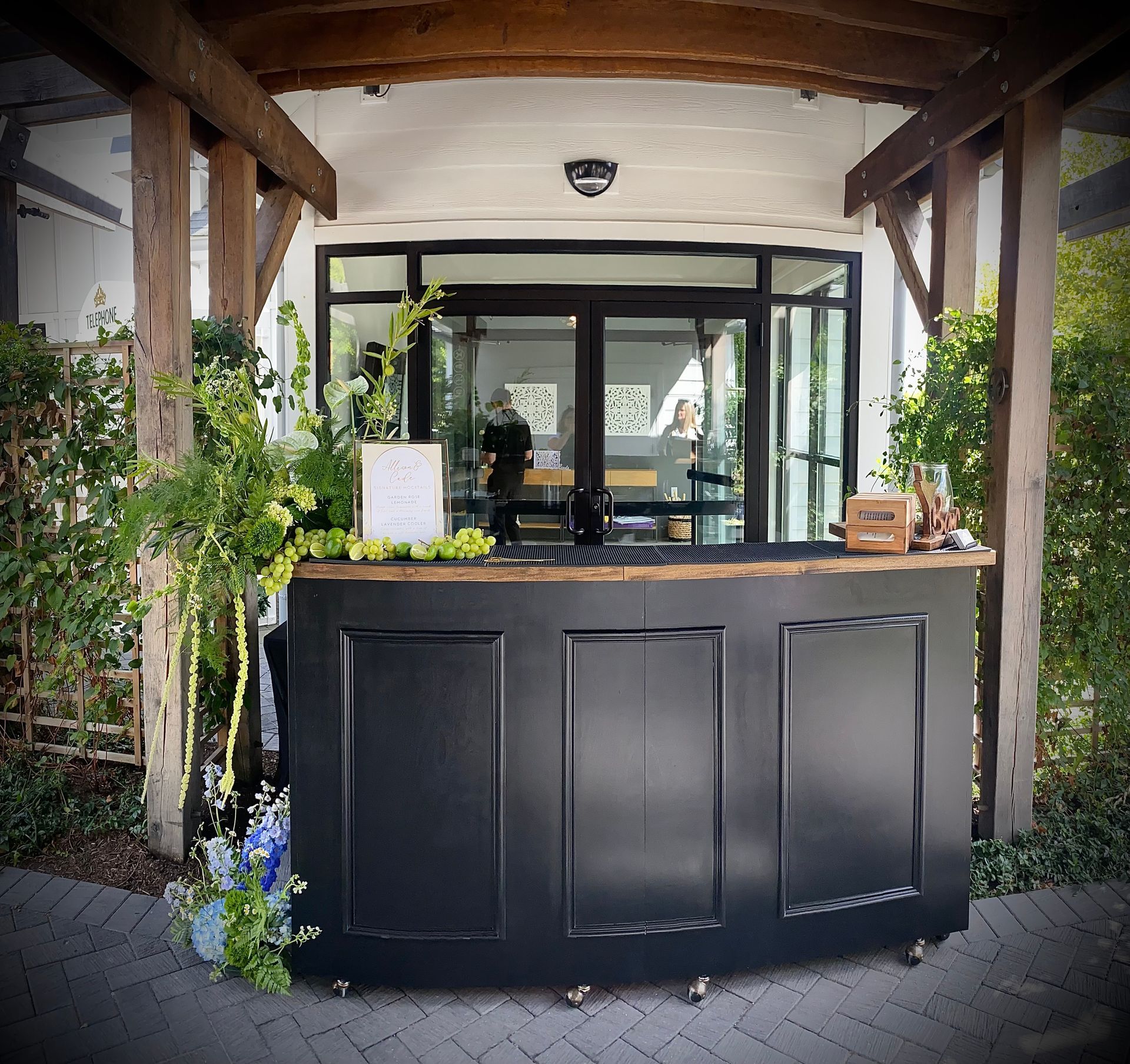 Bartender pours drink from shaker into a glass on a bar; tropical setting, sunset in background.