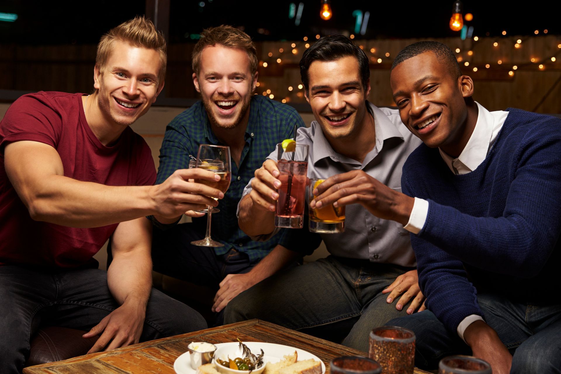 Four diverse men smiling, toasting drinks in a bar, enjoying a social gathering.