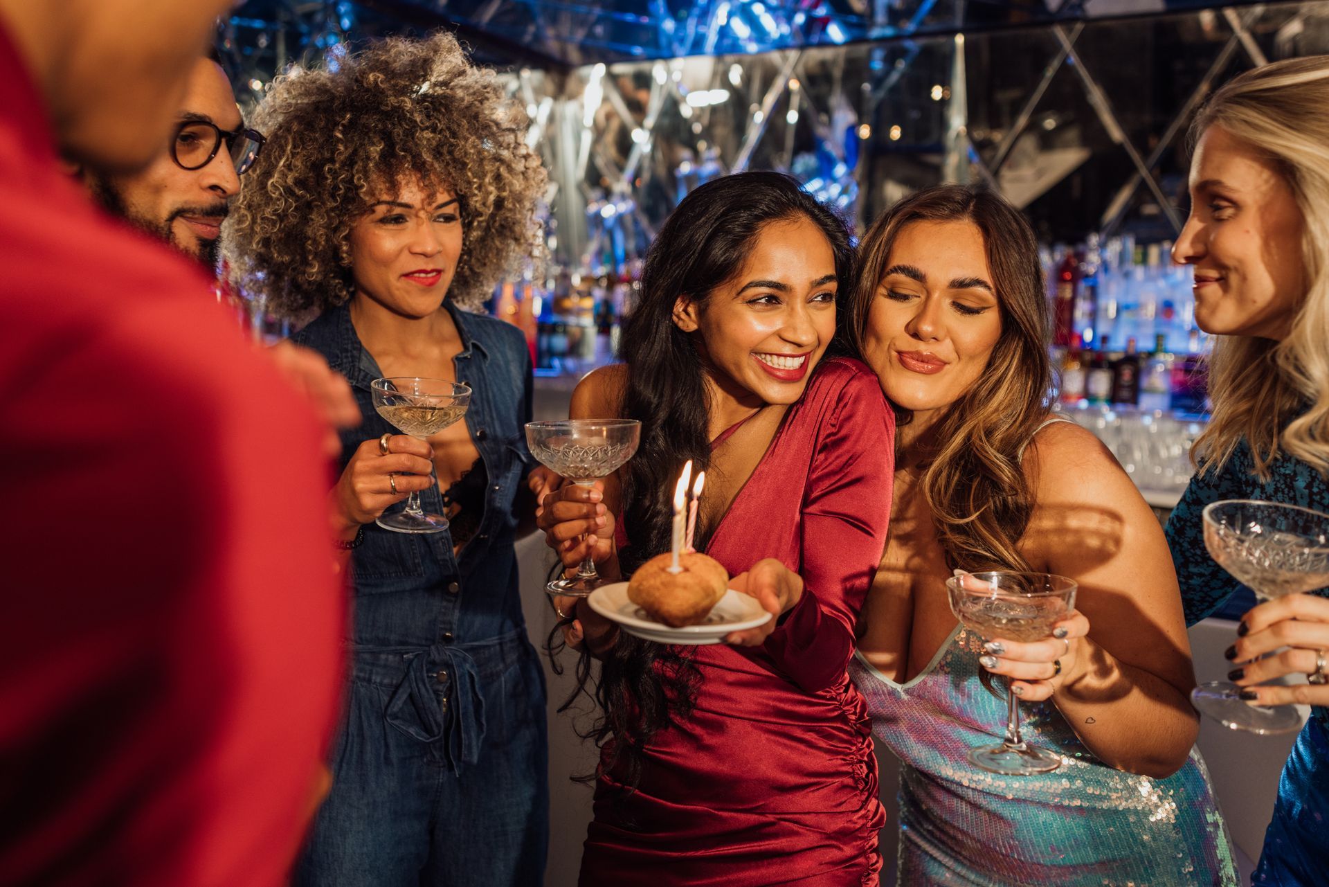 Group of friends celebrating with drinks. Woman holding cake with a candle. Bar setting with bright lights.