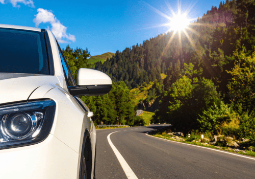 White SUV driving along a winding mountain road bordered by trees under bright summer sunshine.