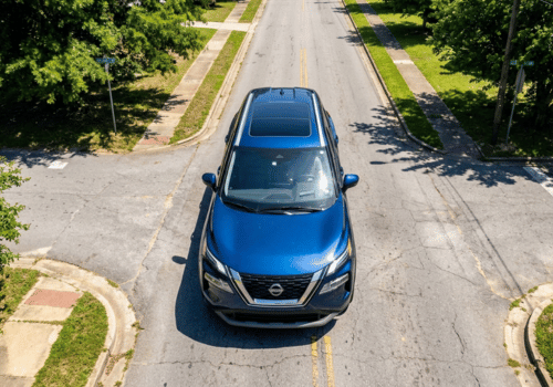 Aerial view of a blue SUV driving through a quiet residential neighborhood with tree-lined streets on a sunny day.