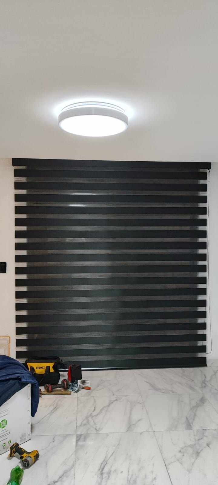 Persianas de rayas blancas y negras sobre una pared blanca, con una lámpara de techo brillante encima. Suelo de mármol.