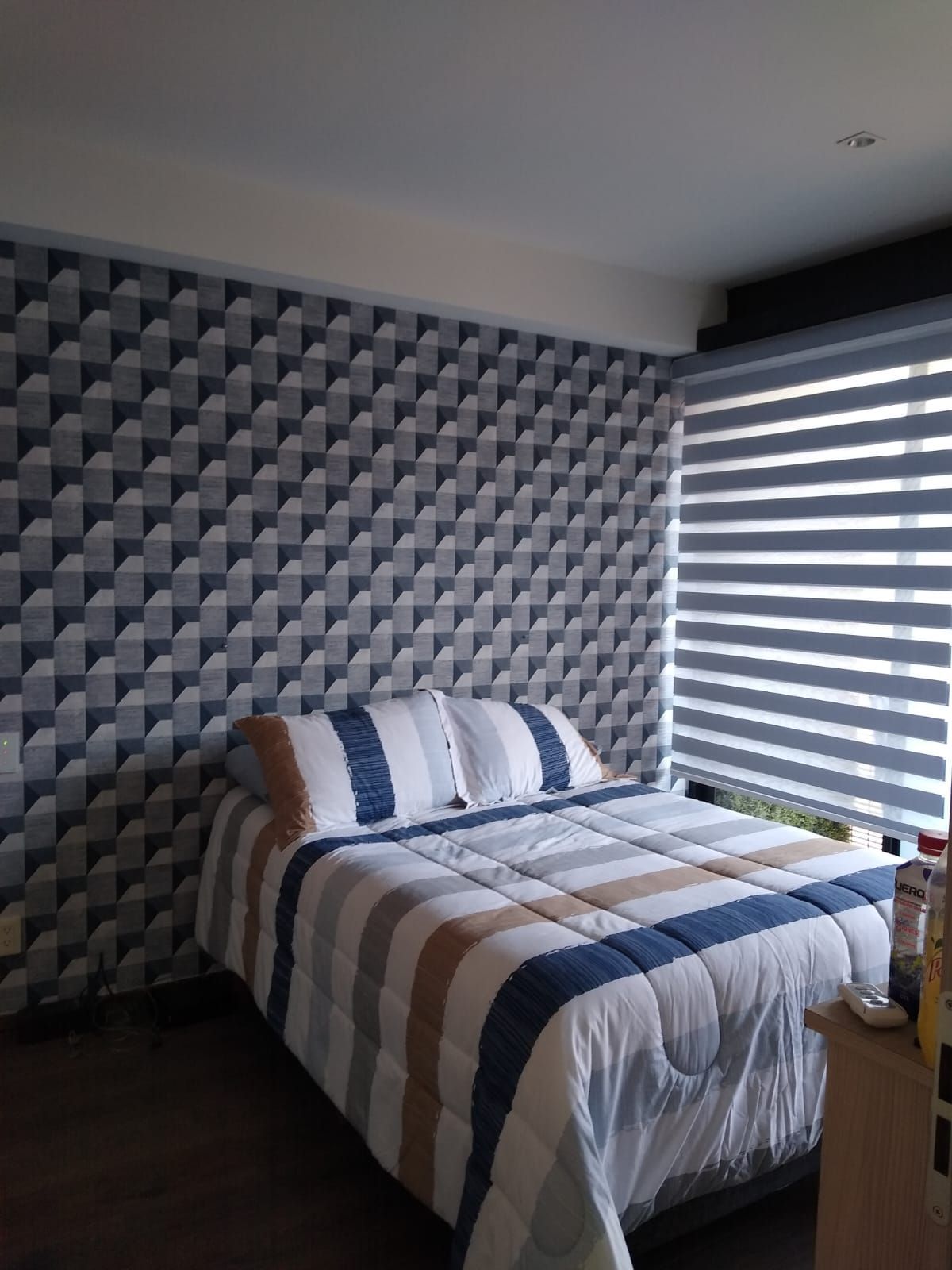 Dormitorio con papel pintado estampado, cama con ropa de cama de rayas y una ventana con persianas.