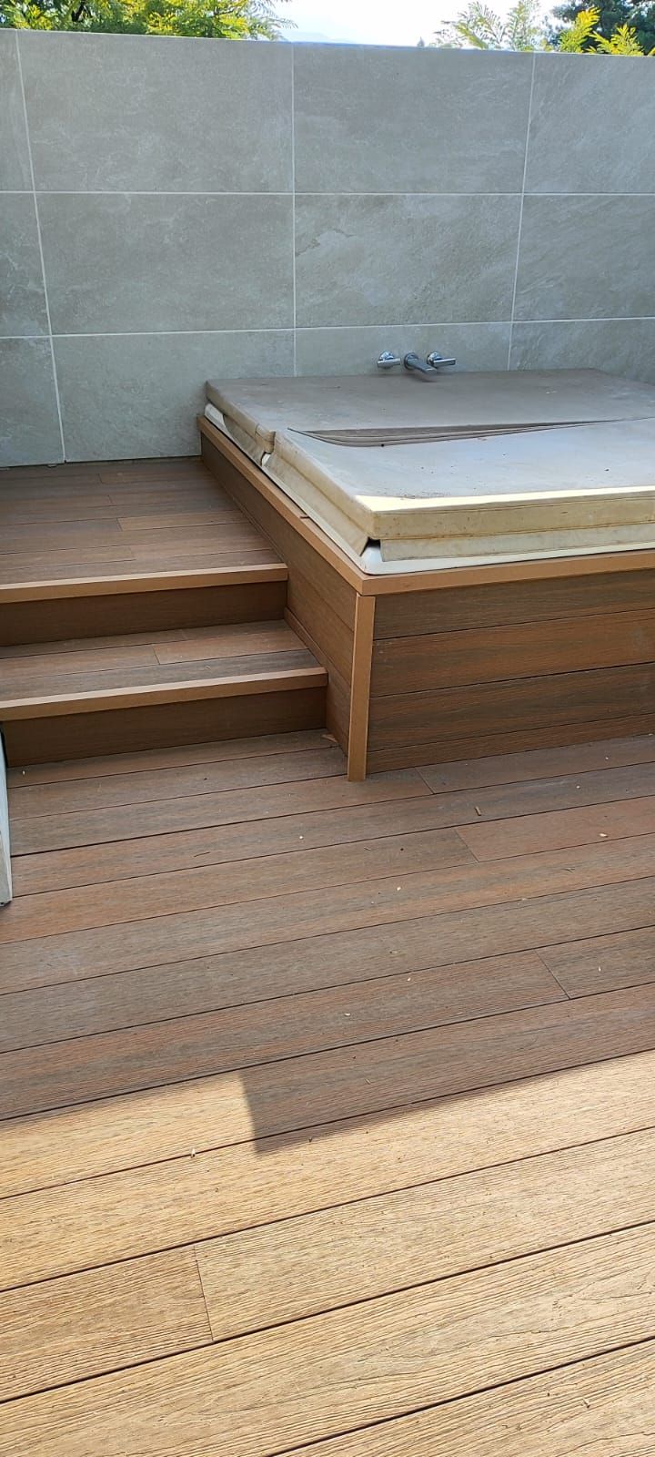 Terraza de madera con escalones que conducen a un jacuzzi elevado contra una pared de azulejos, al aire libre.