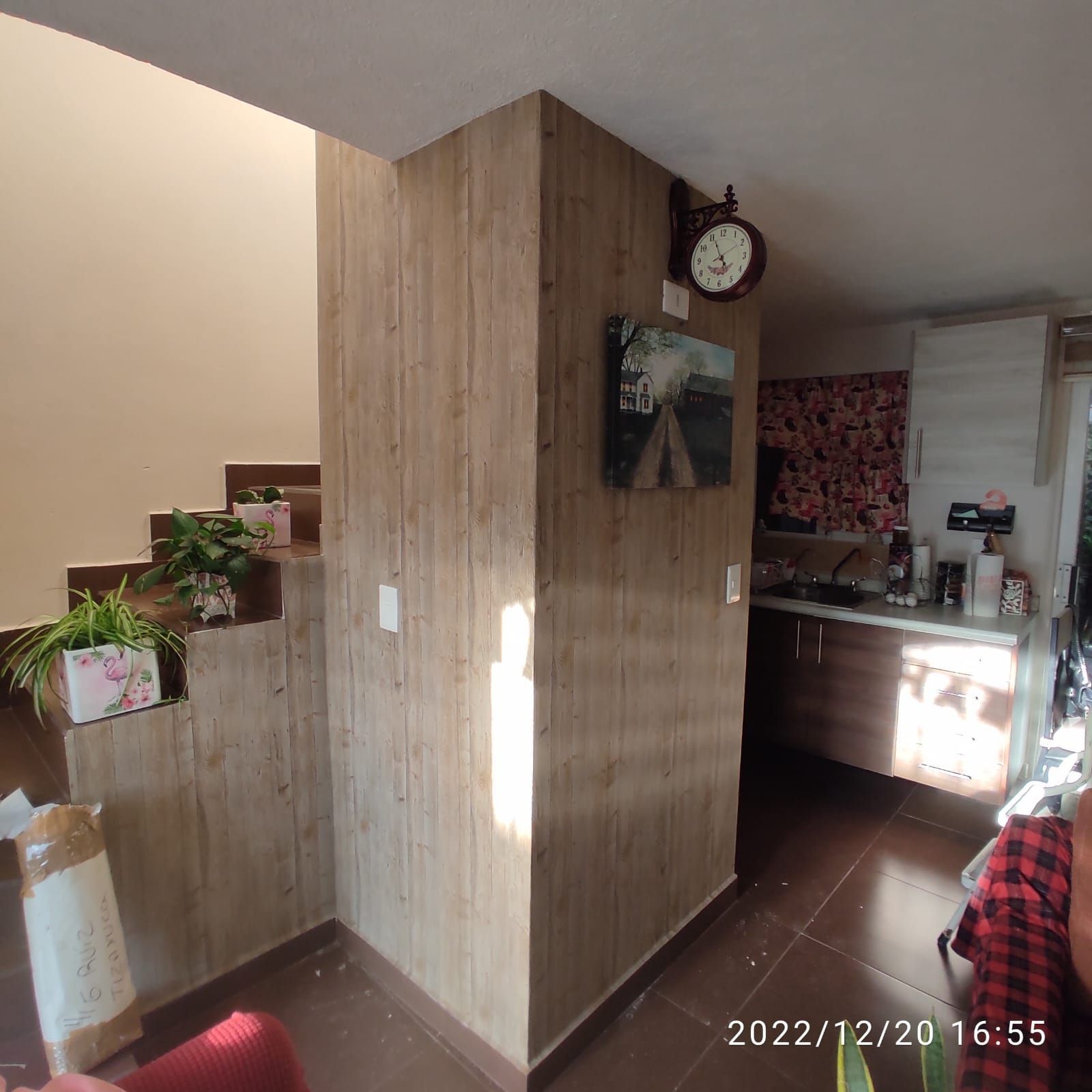 Interior con pared revestida de madera, escalera y reloj. Cocina con armarios a la vista. Marrón, tostado y blanco.