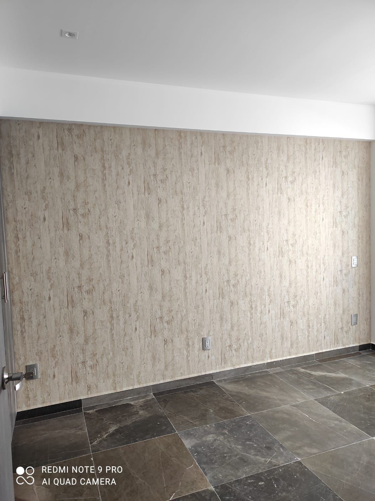 Habitación vacía con piso de baldosas de piedra, pared texturizada de color beige y techo blanco.