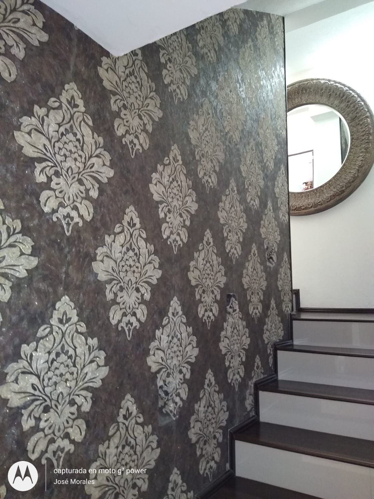 Pared con papel pintado damasco junto a una escalera y un espejo redondo.