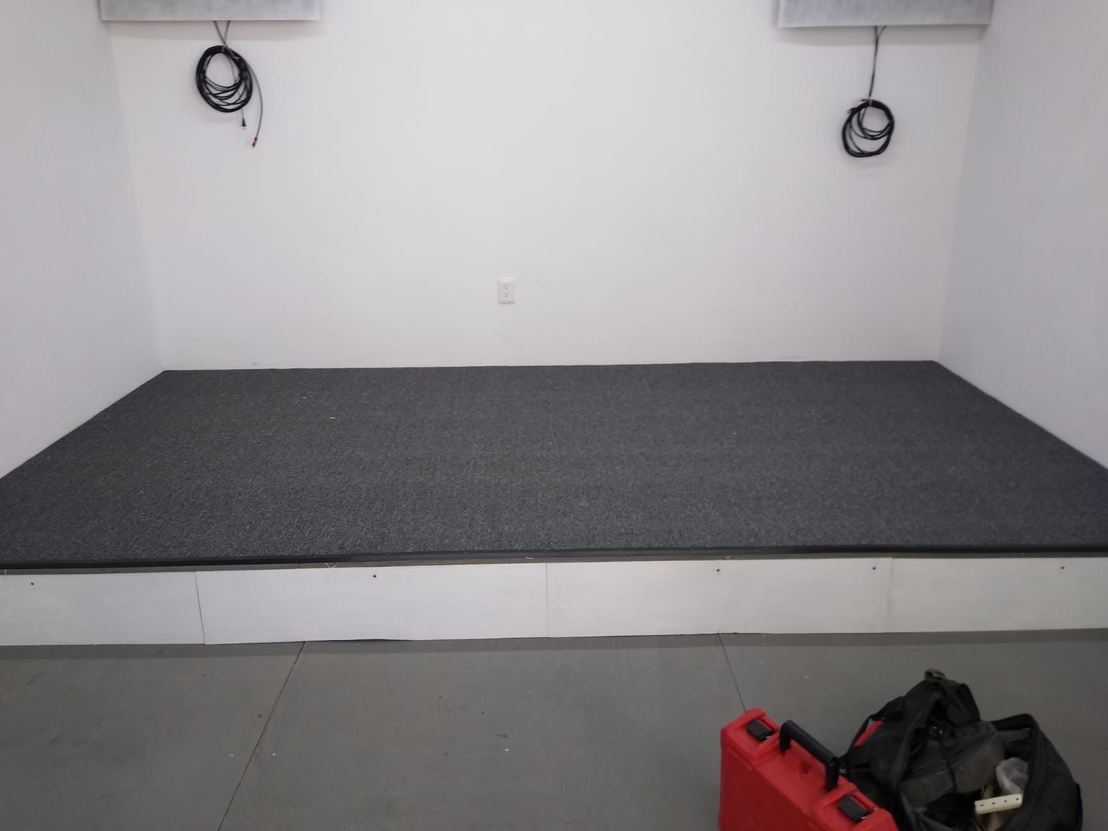 Un pequeño escenario con alfombra gris contra una pared blanca. Cables y un contenedor rojo en el fondo.