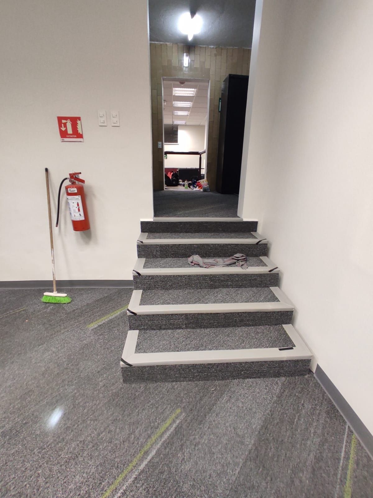 Escalera que conduce a un pasillo con extintor, escoba y suelo gris.
