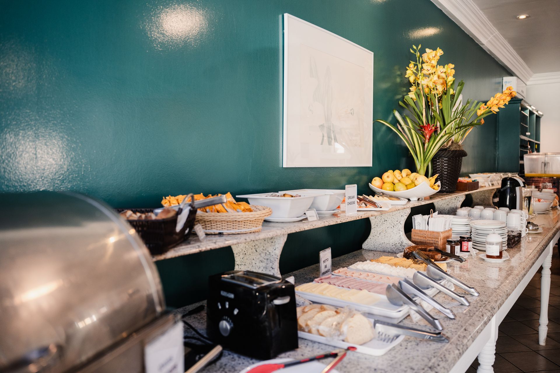 Buffet com diversos alimentos: doces, frutas, carnes e condimentos em um balcão comprido contra uma parede verde-azulada.