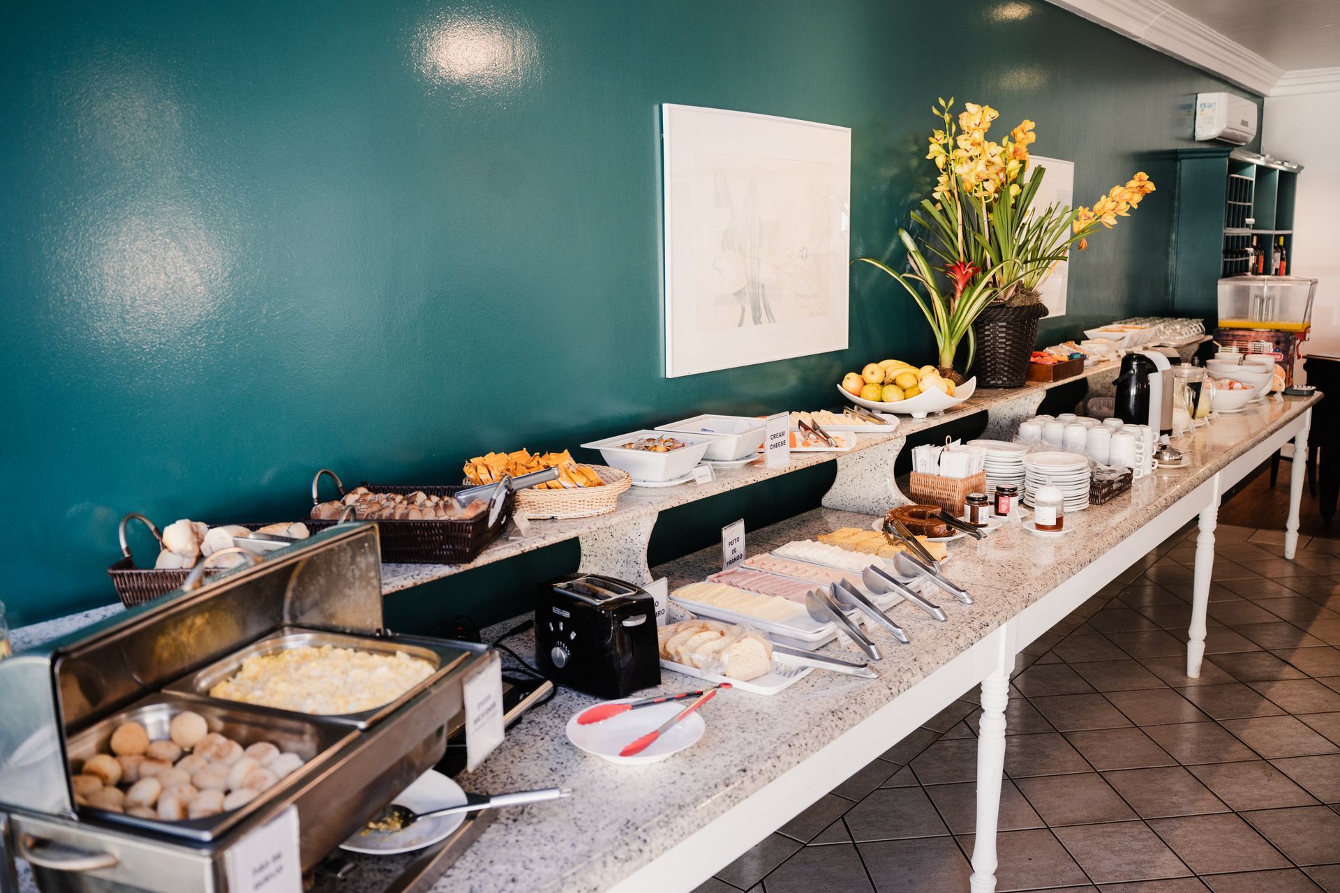 Mesa de buffet posta com diversos itens de café da manhã contra uma parede verde-azulada.