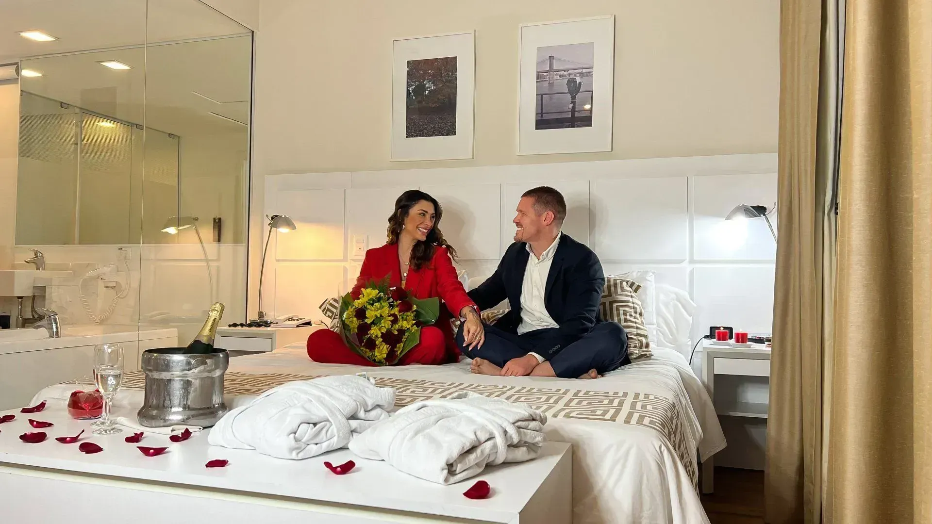 Um casal sentado na cama com buquê de rosas, 2 roupões de banho, pétalas de rosas e uma garrafa de espumante
