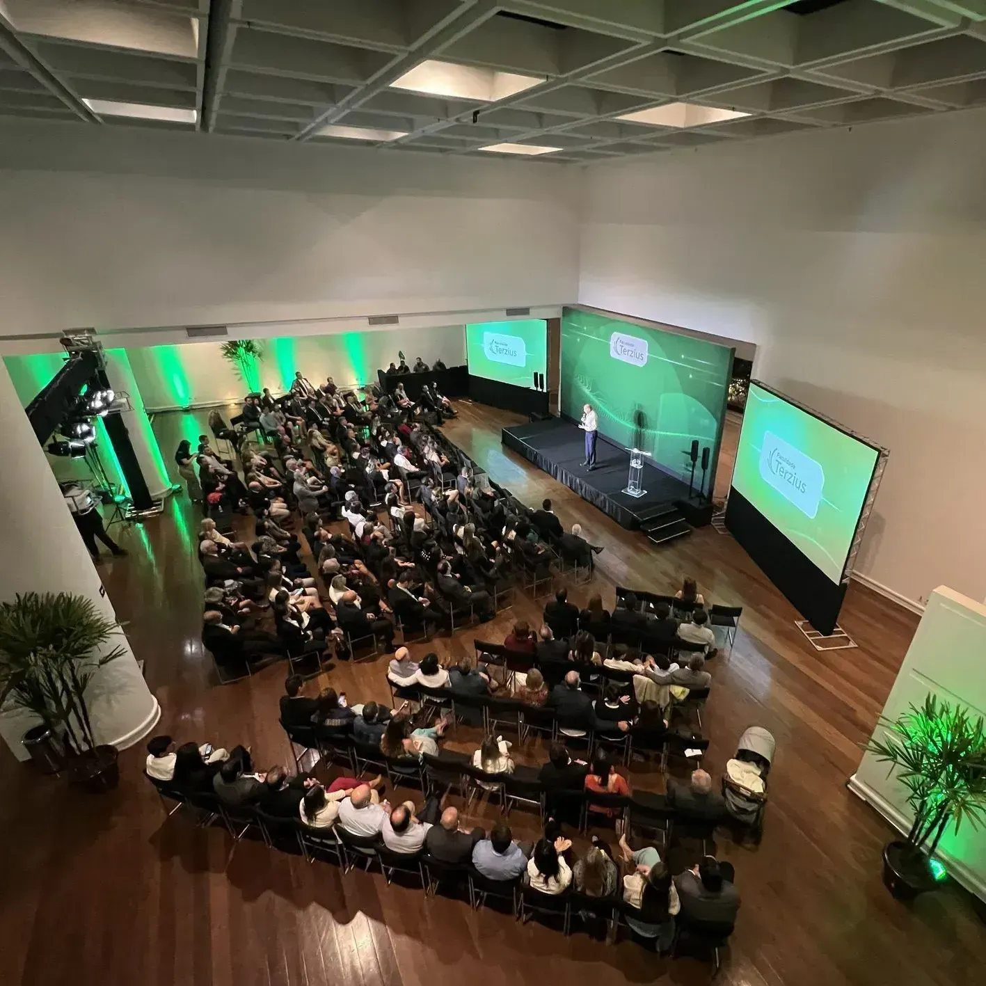 Público em sala de conferências com apresentador no palco, detalhes em verde, duas telas, vista aérea.