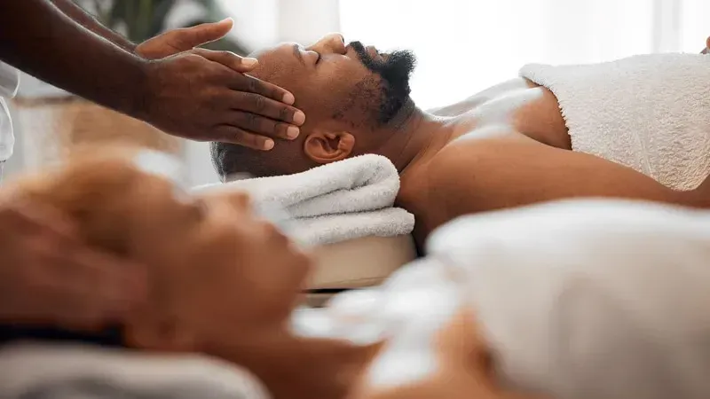 Pessoa recebendo uma massagem facial em um spa. Mãos no rosto, toalhas, ambiente interno.