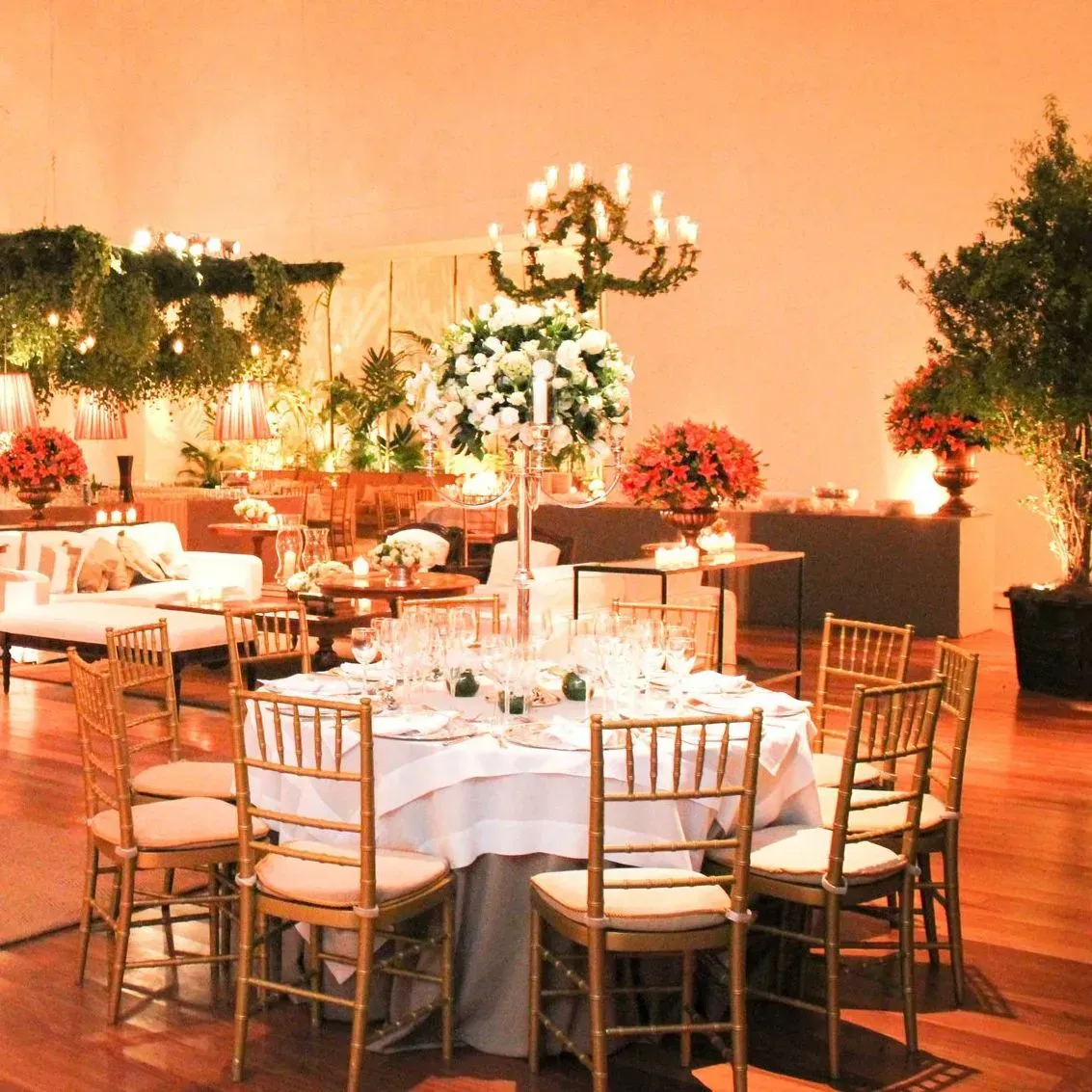 Espaço elegante para eventos, mesa redonda posta para jantar com cadeiras douradas. Arranjos florais amplos e iluminação suave.