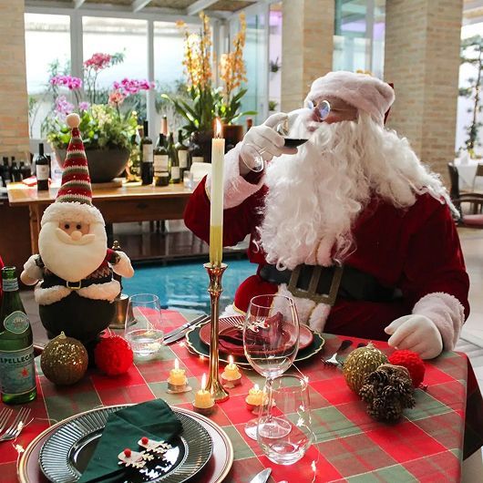 Papai Noel bebendo vinho em uma mesa festiva com um pequeno boneco de Papai Noel, velas e garrafas de vinho ao fundo.