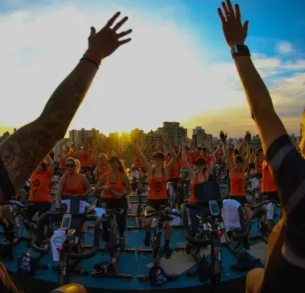 Pessoas em bicicletas ergométricas com os braços erguidos diante de um pôr do sol ao ar livre.