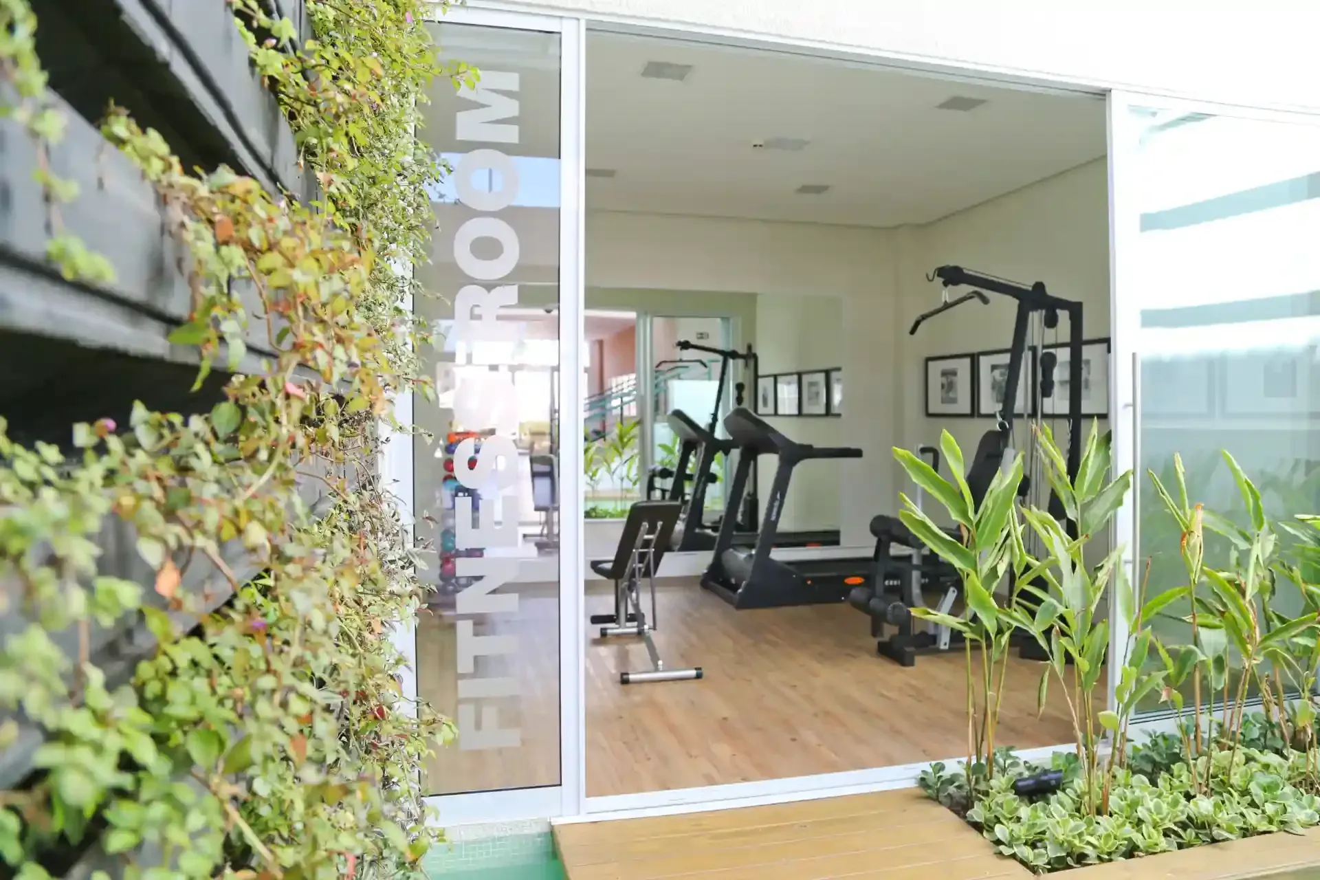 Sala de ginástica com equipamentos de exercício, visível através de portas de vidro. A entrada é cercada por vegetação.