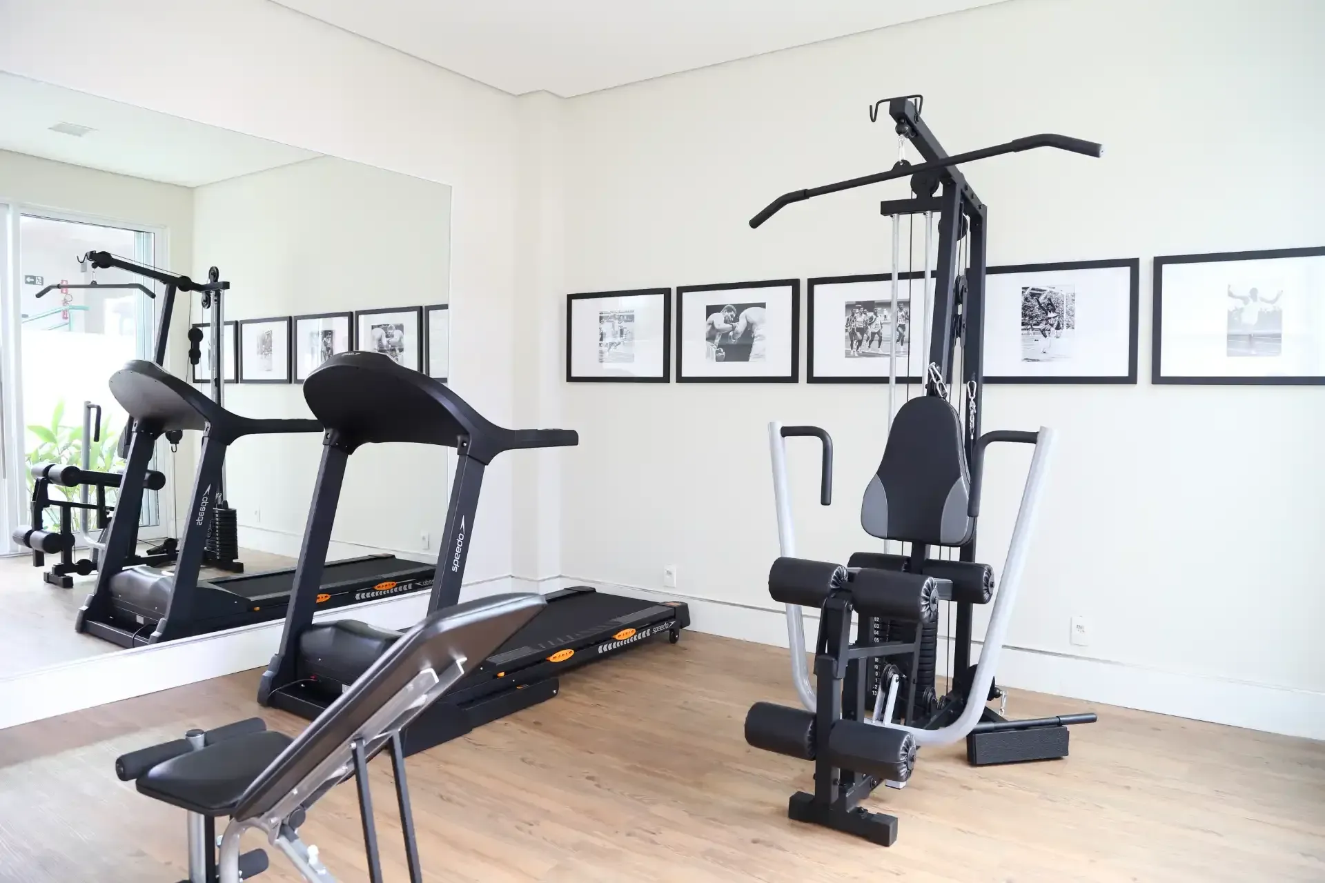 Academia em casa com esteira, aparelho de musculação e obras de arte emolduradas em paredes brancas.