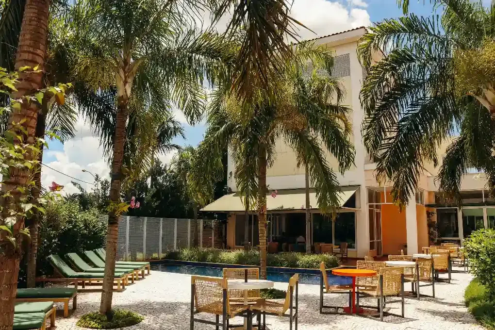 Exterior do hotel com piscina, área de estar externa, palmeiras e céu azul.